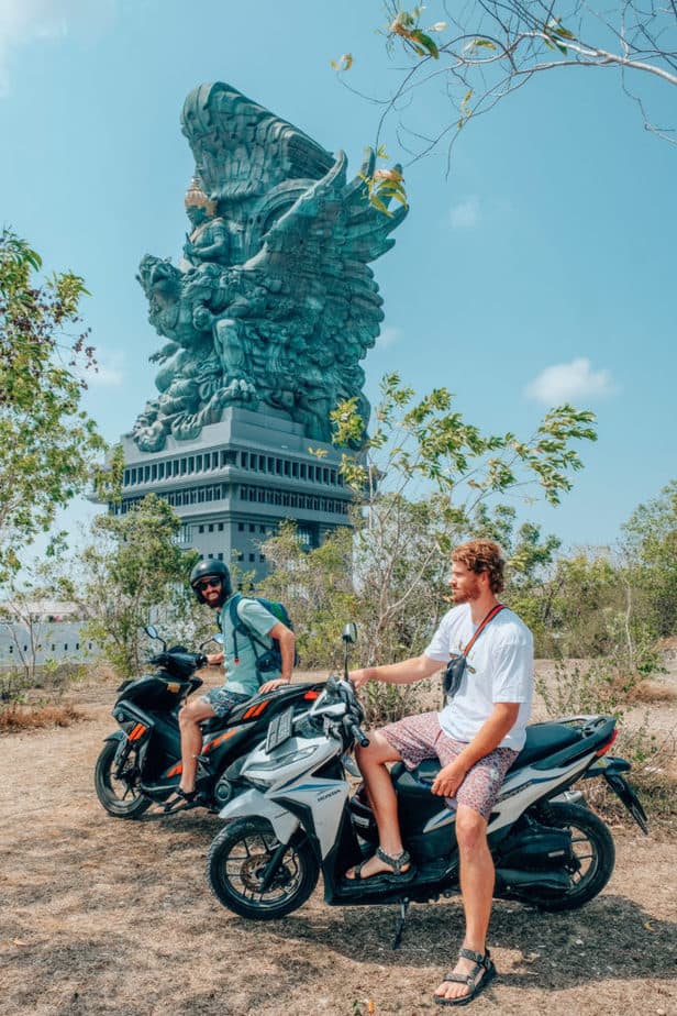 Two men riding scooters in Bali in front of a giant statue