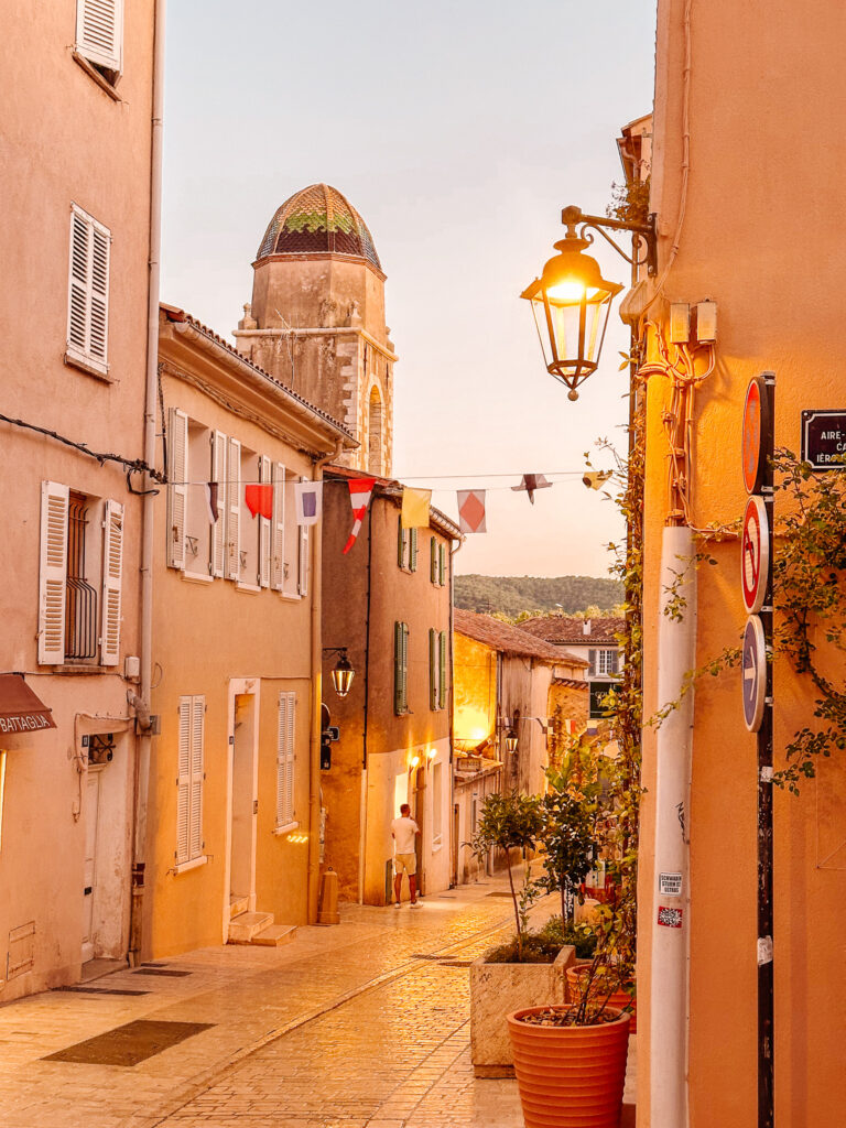 A golden-lit street in Saint-Tropez at dusk, with a bell tower behind it